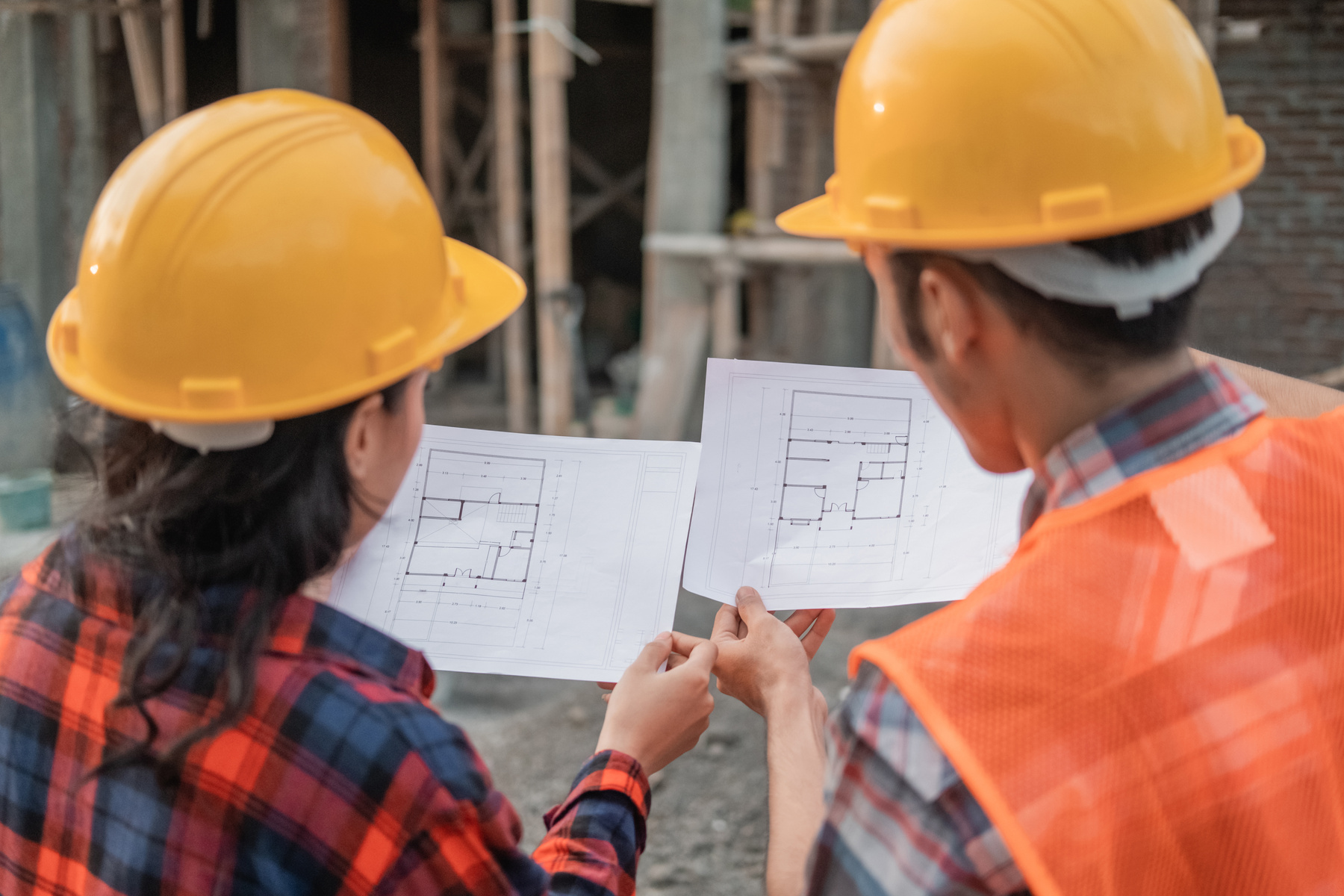 View from behind of Two Asian Contractors Standing in Safety Helmets Holding the Site Plan Building